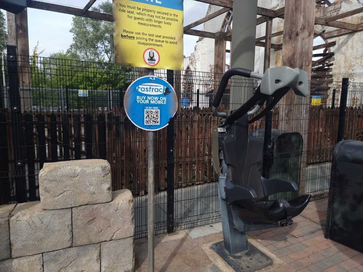 A dark grey rollercoaster test seat with sturdy harnesses sits near a Fastrac QR sign and a warning about body size limitations. This setup assists with managing expectations for guests, especially those concerned about fit, highlighting the importance of checking ahead when navigating plus size at Thorpe Park.