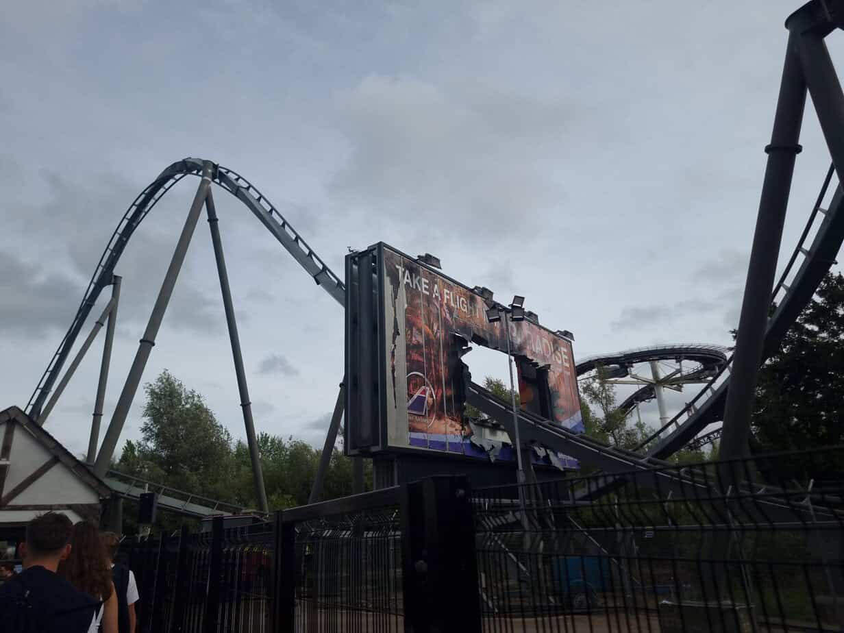 A rollercoaster track dives through a ripped billboard that reads “Take a flight to paradise,” with looping grey track and guests entering the queue in the foreground.