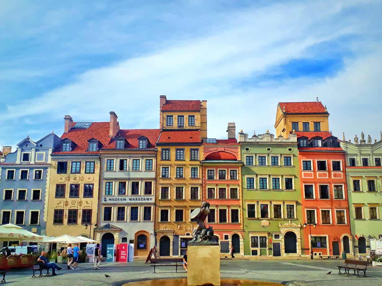 Row of colorful Old Town buildings with ornate facades and red-tiled roofs behind a central fountain — Lively historic charm surrounding the Museum of Warsaw.