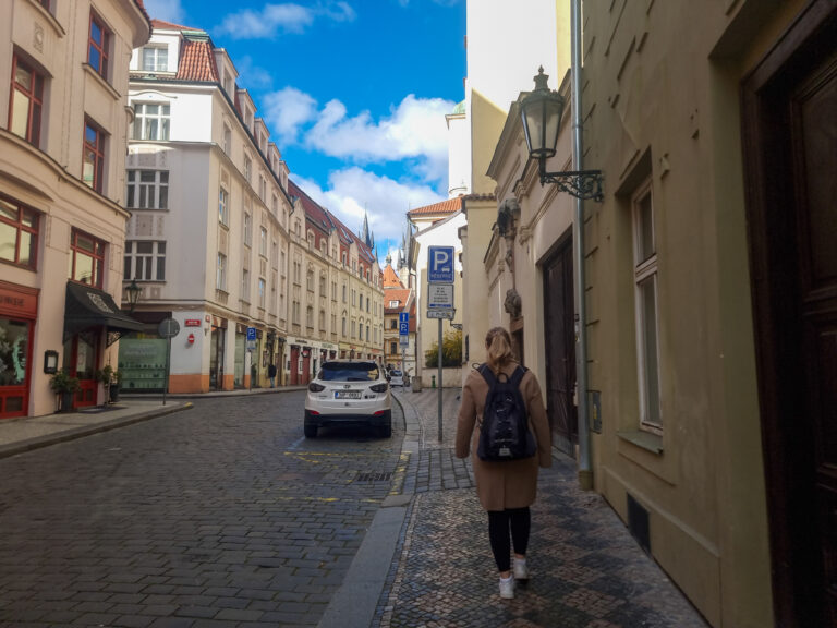 A woman with a tan coat and black backpack walks down a quiet cobblestone street in Prague’s Old Town, framed by pastel buildings and vintage street lamps under a clear blue sky.