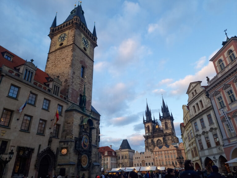 The Old Town Hall Tower with its astronomical clock stands prominently under a blue sky at Prague’s Old Town Square, with the Church of Our Lady before Týn in the background. A bustling crowd adds life to this historic Gothic landmark.