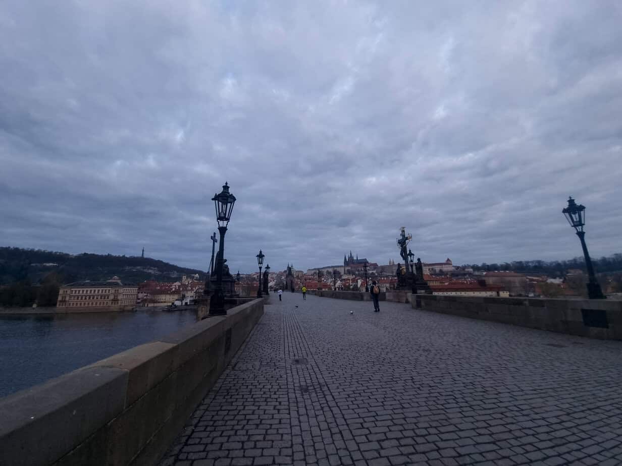 The Charles Bridge at dusk with only a few people walking along its cobblestone path, flanked by statues and lampposts, with Prague Castle in the distance under a moody sky.
