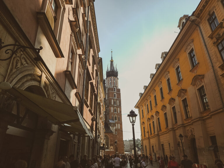 A narrow Kraków street lined with historic townhouses leads to the iconic twin towers of St. Mary’s Basilica, glowing in late afternoon light.
