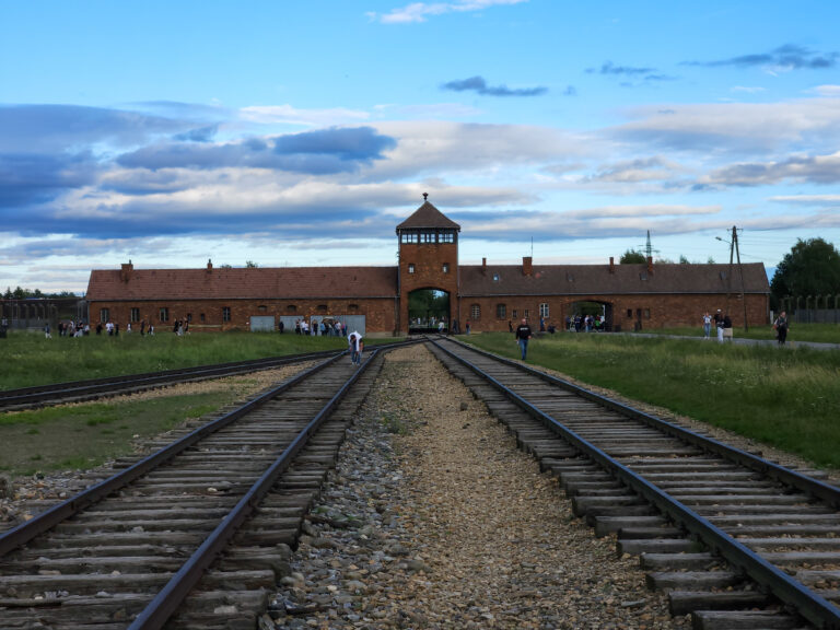 View of the railway tracks leading to the brick gatehouse of Auschwitz II-Birkenau under a partly cloudy sky, with visitors walking along the tracks toward the entrance.