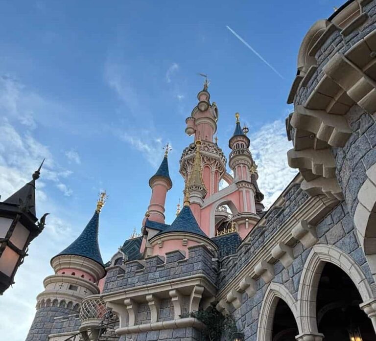 Close up view of a pink fairytale castle with blue rooftops and ornate golden details against a partly cloudy sky. Stone archways and turrets frame the scene, highlighting the iconic Disneyland Paris architecture.