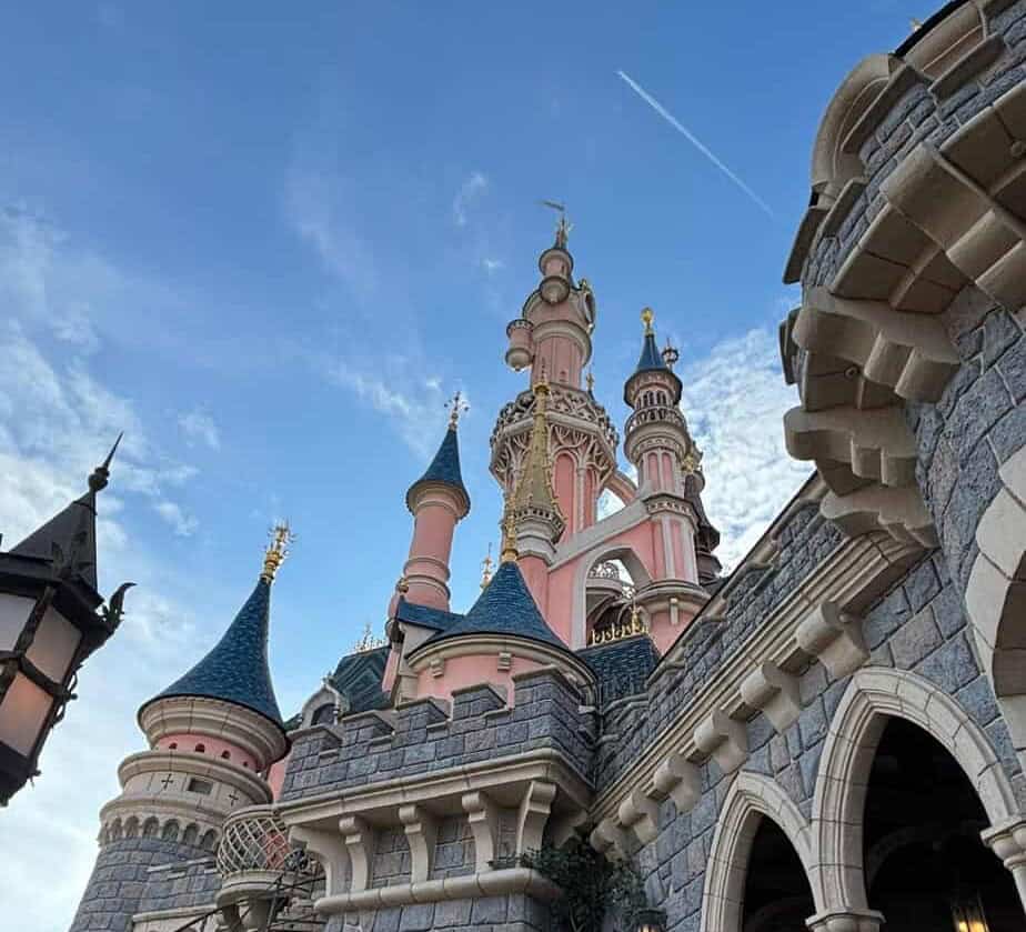 Close up view of a pink fairytale castle with blue rooftops and ornate golden details against a partly cloudy sky. Stone archways and turrets frame the scene, highlighting the iconic Disneyland Paris architecture.