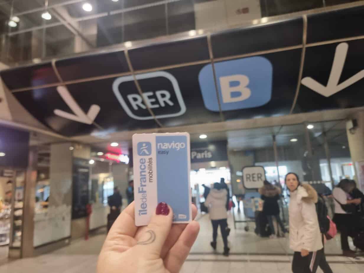 A hand with red nails holds up an RER B ticket under directional signage. This shows one of the cheaper ways to get from Charles de Gaulle Airport to Disneyland Paris.