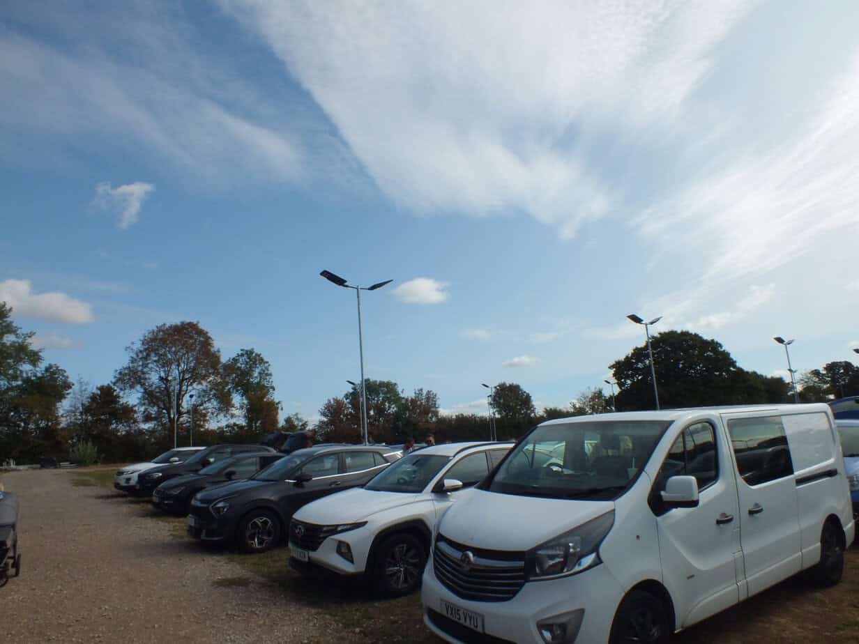 Rows of cars and a white van parked in the outdoor car park at Twycross Zoo beneath a blue and cloudy sky.