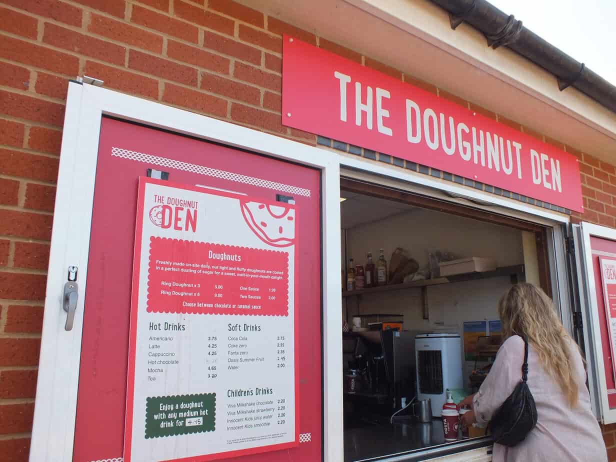 The Doughnut Den food kiosk at Twycross Zoo with a customer ordering at the serving window. A large red menu beside the window lists doughnuts along with hot drinks soft drinks and childrens drinks.