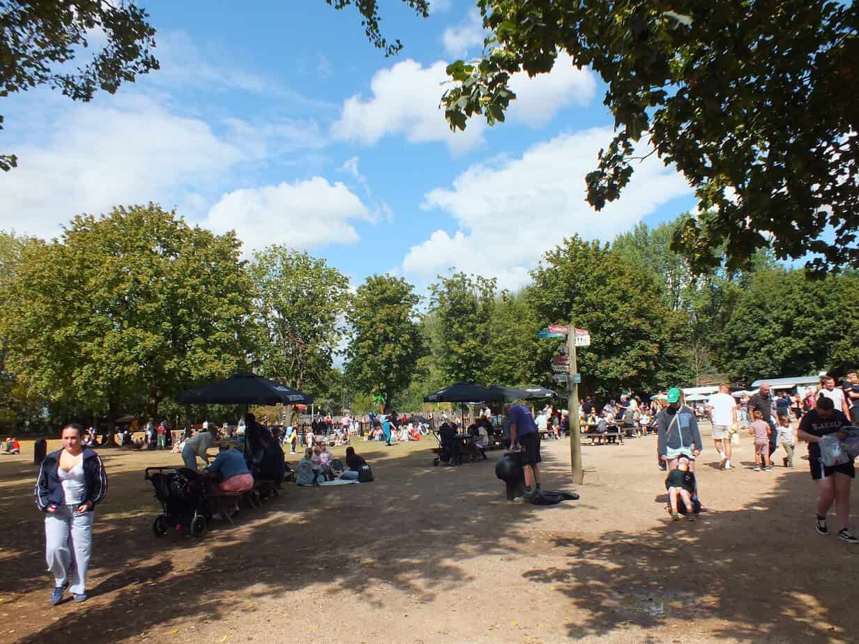 Busy open area at Twycross Zoo surrounded by leafy trees with families walking resting at picnic tables and pushing strollers. Large umbrellas provide shade around the seating areas on a sunny day.